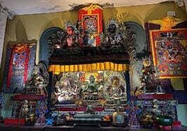 A home altar   on a side cabinet in the dining room with the Three Supremes, Tara, Ushnisha Vijaya and Vajrasattva in the center, lit with strip lights, incense in front, flanked by the eight auspicious symbols as offeirngs. On top of the main altar is a shelf raising up two other aspects of Tara (Red and black) along with Vajrakilaya. To the right is yellow Tara and a thangka of Tara's wrathful form Palden Lhamo. To the left is the great Heruka of the Padma Family Hayagriva on a Thangka and a statue of Jambhala.