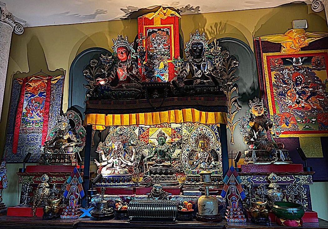 A home altar   on a side cabinet in the dining room with the Three Supremes, Tara, Ushnisha Vijaya and Vajrasattva in the center, lit with strip lights, incense in front, flanked by the eight auspicious symbols as offeirngs. On top of the main altar is a shelf raising up two other aspects of Tara (Red and black) along with Vajrakilaya. To the right is yellow Tara and a thangka of Tara's wrathful form Palden Lhamo. To the left is the great Heruka of the Padma Family Hayagriva on a Thangka and a statue of Jambhala.